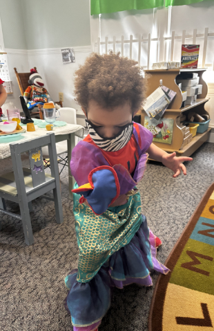 A child in a daycare dressed up in colorful clothes with a puppet on his hand.
